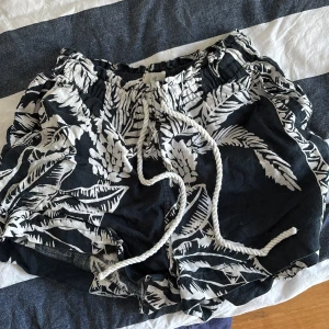 Svartvita blommiga shorts från H&M - Säljer ett par snygga svartvita shorts från H&M med blommigt mönster. De har en bekväm elastisk midja och snörning framtill. Perfekta för sommaren.