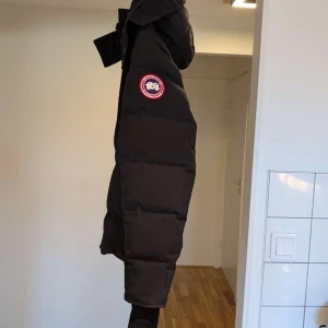 Svart dunjacka från Canada Goose - Säljer en svart dunjacka från Canada Goose med huva och märkets logga på ärmen. Jackan har långa ärmar och stängs med dragkedja och knappar. Perfekt för kalla vinterdagar.