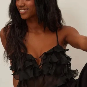 Svart volangprydd topp från NA-KD - Säljer en elegant svart topp från NA-KD med tunna axelband och flera lager av volanger. Toppen är perfekt för en festlig kväll och ger en luftig och stilren look.