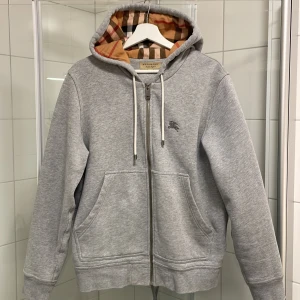 Grå hoodie från Burberry - Säljer en stilren grå hoodie från Burberry med dragkedja och klassiskt rutigt foder i huvan. Tröjan har en broderad logga på bröstet och praktiska fickor framtill. Perfekt för en avslappnad och trendig look. Inköpt på Nordiska Kompaniet, NK. 