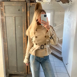 Beige trenchcoat med bälte - Snygg beige trenchcoat med bälte och knappar. Perfekt för vår och höst. Jackan har långa ärmar och en klassisk design som passar till många olika stilar. Aldrig använd!