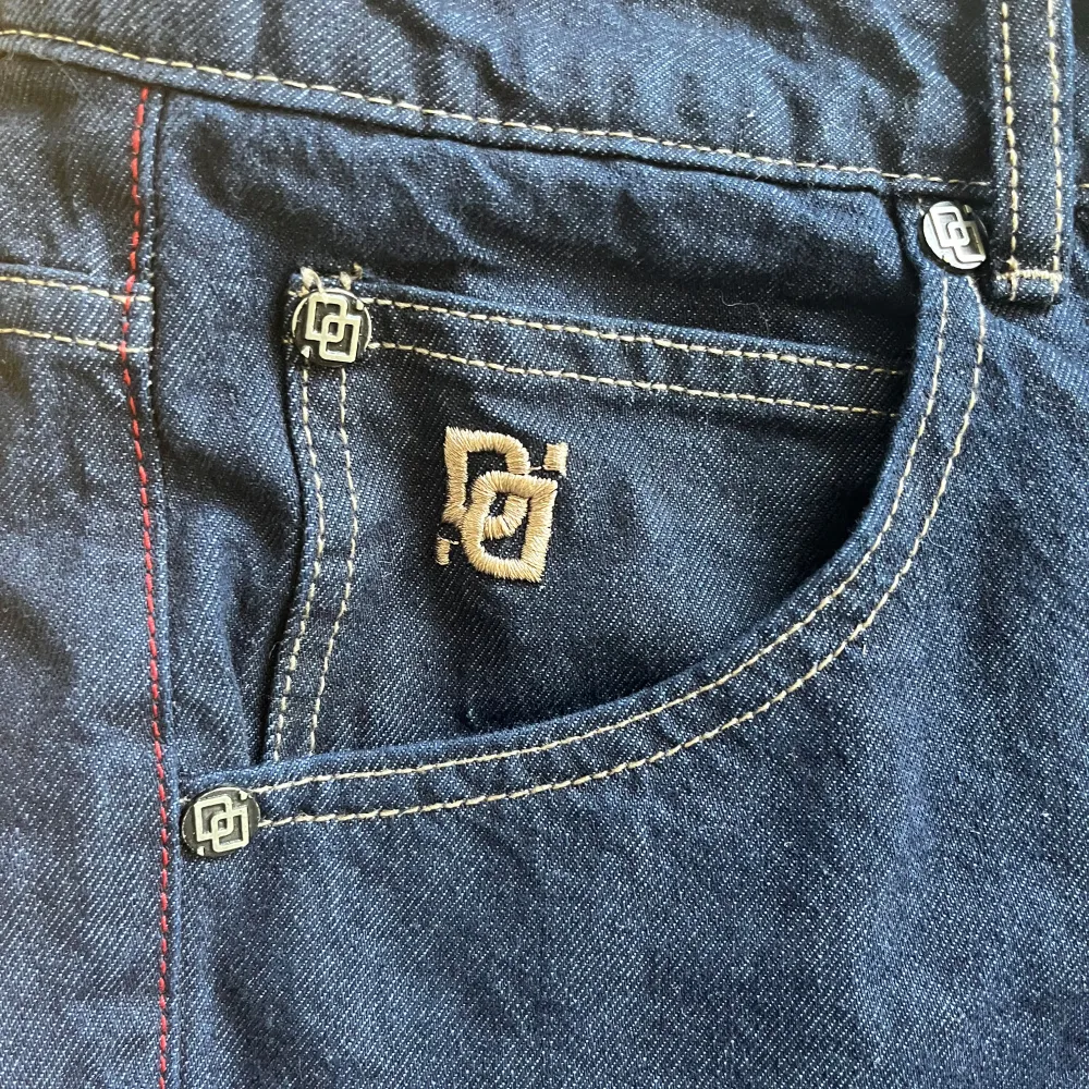 Snygga mörkblå jeans från DNGRS med broderad logga på bakfickan och kontrastsömmar. Klassisk femficksdesign med knappgylf och bälteshällor. Perfekta för en avslappnad stil.. Farkut & Housut.