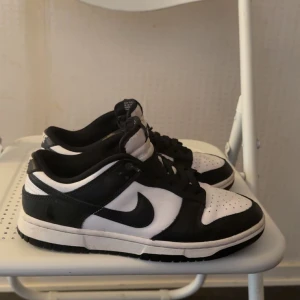 Nike Dunk Low i svart och vitt - Snygga Nike Dunk Low sneakers i klassisk svart och vit färgkombination. Skorna har en låg profil med snörning och den ikoniska Nike-loggan på sidan. Perfekta för en stilren och sportig look.