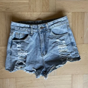Blå jeansshorts med slitningar - Säljer ett par coola blå jeansshorts med slitningar framtill. De har en klassisk femficksdesign och är perfekt för en avslappnad stil. Shortsen har en knapp och dragkedja framtill.
