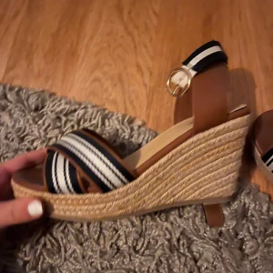 Bruna kilklackar med randigt band - Snygga bruna sandaler med kilklack och randigt band i svart och vitt. Sandalerna har en justerbar rem med spänne runt ankeln och en bekväm sula. Perfekta för sommarens alla tillfällen.