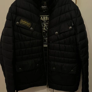 Svart quiltad jacka från Barbour - Säljer en svart quiltad jacka från Barbour International. Jackan har en dragkedja framtill och flera fickor med knappar. Den är perfekt för kyligare dagar och har en stilren design med Barbour-logga på bröstet.