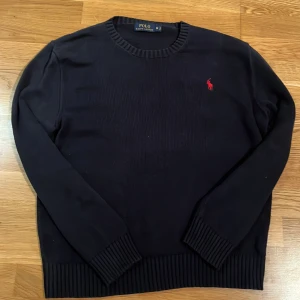 Mörkblå  tröja från Polo Ralph Lauren - Säljer en stilren svart tröja från Polo Ralph Lauren med det klassiska röda logotypbroderiet på bröstet. Tröjan har långa ärmar och ribbade muddar vid ärmslut och nederkant. Perfekt för en elegant och avslappnad look. Skick 9/10! Köpt från plick innan men inte kommit till så mycket användning. Inga defekter.
