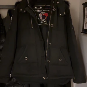 Svart dunjacka från Moose Knuckles - Säljer en svart dunjacka från Moose Knuckles med pälsdetaljer på huvan. Jackan har dragkedja och knappar framtill samt broderad logotyp på insidan. Perfekt för kalla dagar. Den är helt ny och jag har bara använt den i en månad. köpt för 15k
