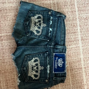 Mörkblå jeansshorts med broderad krona -  Skriv via intresse innan ni köper för att är inte hemma nu kommer hem 5 april så då kommer varan skickas .Snygga mörkblå jeansshorts från Victoria Beckham med fransiga kanter och broderade kronor på bakfickorna. Perfekta för en avslappnad stil med en touch av lyx. De har en klassisk femficksdesign och knappgylf.