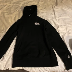 Svart hoodie från Billionaire Boys Club - Säljer en svart hoodie från Billionaire Boys Club med logga på bröstet och en astronautdetalj på ryggen. Tröjan har en klassisk känguruficka och ribbade muddar vid ärmslut och nederkant. Perfekt för en avslappnad stil.