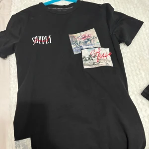 Svart t-shirt från Supply & Demand - Snygg svart t-shirt från Supply & Demand med tryck på bröstet. T-shirten har en cool design med text och grafiska element i rött och vitt. Perfekt för en avslappnad stil. Helt ny endast använd en gång 