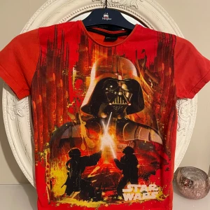 Röd Star Wars t-shirt med Darth Vader - Cool röd t-shirt med ett stort tryck av Darth Vader och en episk ljussabelstrid. Perfekt för alla Star Wars-fans som vill sticka ut. Tillverkad av bekvämt material och har korta ärmar. Perfekt för att visa din kärlek till galaxens mest ikoniska saga!vintage .storlek xs .kan möta er i Stockholm utanför Åhléns i T-Central