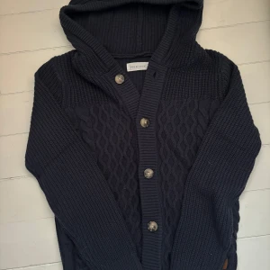 Mörkblå stickad kofta från Pier One - Säljer en snygg mörkblå stickad kofta från Pier One med huva och knappar framtill. Koftan har ett flätmönster och ribbade detaljer vid ärmslut och nederkant. Perfekt för kyliga dagar!