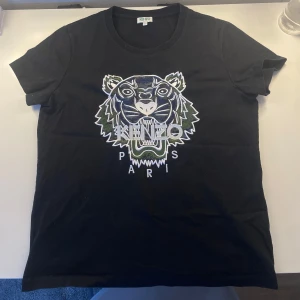 Svart t-shirt med tigertryck från Kenzo - Säljer en svart t-shirt från Kenzo med ett coolt tigertryck i vitt och grönt. T-shirten har korta ärmar och en normal passform. Perfekt för dig som vill ha en stilren men ändå iögonfallande look. 🐯