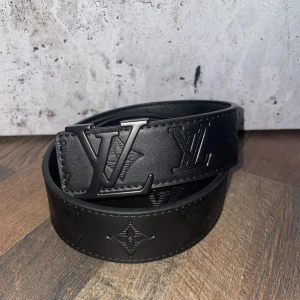 Svart bälte från Louis Vuitton - Snyggt svart bälte från Louis Vuitton med ett stilrent LV-spänne. Bältet är tillverkat i skinn och har en diskret präglad design längs remmen. Perfekt för att ge en lyxig touch till din outfit.