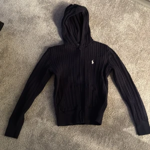 Svart stickad hoodie från Ralph Lauren - Säljer en snygg svart stickad hoodie från Ralph Lauren med dragkedja och det klassiska logotypbroderiet på bröstet. Tröjan har långa ärmar och en mysig huva, perfekt för kyliga dagar. Passar både till vardags och mer avslappnade tillfällen.