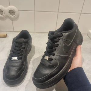 Svarta Air Forces - Snygga svarta Nike Air Forces med klassisk design och snörning. Perfekta för en stilren look. Skorna har en robust sula och ikonisk Nike-logga på sidan.