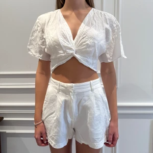 Vit croppad topp från Zara - Säljer en stilren vit croppad topp från Zara. Blusen har korta ärmar och en knytning framtill som ger en snygg detalj. Perfekt för varma dagar och kan enkelt matchas med olika outfits. Tröjan har ett litet hål under vänstra armen, men det är inget som syns tydligt då armen täcker det. Se sista bilden. 