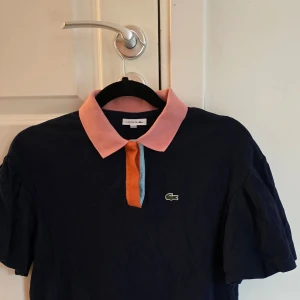Mörkblå pikéklänning från Lacoste - Säljer en snygg mörkblå pikétröja från Lacoste med en kontrasterande rosa krage och orange knappslå. Perfekt för en stilren och avslappnad look. Den har korta ärmar och det klassiska Lacoste-märket på bröstet. Använd 2 ggr st 140. Ny pris 950:-