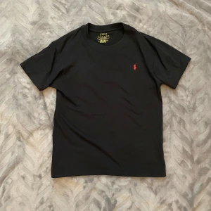 Ralph Lauren T-Shirt - Svart T-shirt från Ralph Lauren med en röd logga på bröstet. Tröjan är använd 2 gånger men har inga som helst tecken på användning eller defekter. 