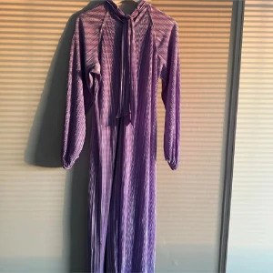 Lång lila plisserad klänning med knyt - Långklänning i plisserat lila tyg med subtila ränder. Standard storlek. Nyskick. Köpt för 400kr