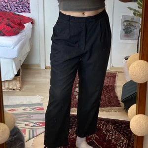 Svarta kostymbyxor med hög midja, M - Snygga svarta kostymbyxor med bra passform. Hög midja och raka, lite vidare ben. Byxorna har pressveck framtill, sidfickor och en bakficka med knapp. Kostymbyxorna är lätt randiga med grått och svart.