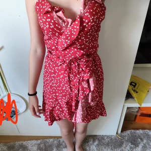 Röd omlottklänning med hjärtan - Supersöt röd kort omlottklänning med vita hjärtan, volangkant och knytband i midjan. Klänningen har smala axelband och en lätt v-ringning framtill. Perfekt för sommarens alla tillfällen!