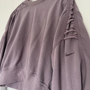 Ljuslila Nike sweatshirt med snörning - Nike Dri-Fit sweatshirt i ljuslila med snygg snörningsdetalj längs axlarna och ärmarna. Jättebra att ha när man tränar eller bara i vardagen!