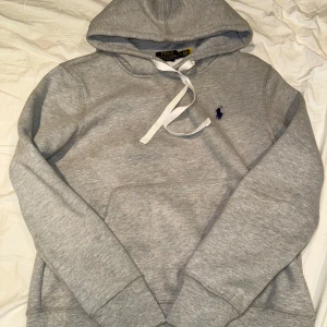 Grå hoodie från Polo Ralph Lauren - Klassisk grå hoodie från Polo Ralph Lauren med vit snörning i huvan och mörkblå broderad logga på bröstet. Tröjan har lång ärm, känguruficka och är tillverkad i mjuk bomullsmix. Perfekt för en chill och stilren look.