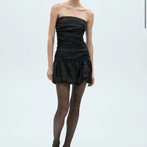 Svart axelbandslös kortklänning - Supersnygg svart axelbandslös kortklänning med tight överdel och puffig kjol. Klänningen har en stilren, minimalistisk look och är perfekt för dig som vill sticka ut. Materialet har en lätt glans och ger en lyxig känsla.