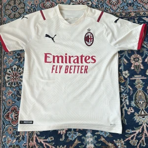 AC Milan Tröja - AC Milan bortatröja i creme vit med röda detaljer och klubbmärke på bröstet. Puma-logga på axlarna och bröstet, samt Emirates Fly Better-tryck på framsidan. Tillverkad i lätt och ventilerande DryCell-material, perfekt för sommaren som är här nu. Den är storlek 13-14 år. Tröjan är snålt använd vilket gör att den i väldigt fint skick. 