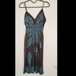 Brun och turkos sommarklänning M/L - Snygg brun klänning med tunna axelband och v-ringning. Klänningen har ett stort turkost blommönster framtill och nertill, vilket ger en bohemisk vibe. Lätt och luftig modell, perfekt för varma dagar. Midjan är smockad för en bekväm passform.