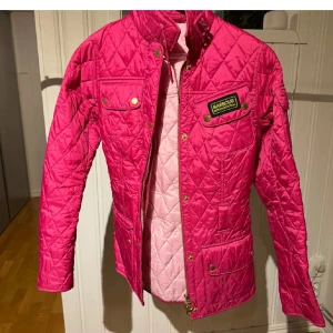 Quiltad rosa jacka från Barbour - Säljer en snygg quiltad jacka från Barbour i stark rosa färg med guldiga knappar och dragkedja. Jackan har flera fickor framtill, Barbour-logga på bröstet och hög krage. Perfekt för dig som vill sticka ut med färg i vår eller höst!