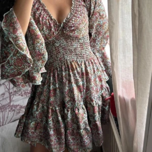 Blommig kortklänning med volangärm - Supersöt kortklänning med blommigt mönster i vinrött, grönt och beige. Klänningen har v-ringning, smockad midja och vida volangärmar. Tyget är lätt och flowy, perfekt för varma dagar. Romantisk och bohemisk vibe!