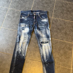 Dsquared2 Skater Jean med slitningar - Snygga blå Dsquared2 Skater Jeans med coola slitningar, färgstänk och trasiga detaljer på benen. Klassisk femficksmodell med smal passform och läderpatch bak. Perfekta för dig som gillar streetstyle och vill sticka ut.