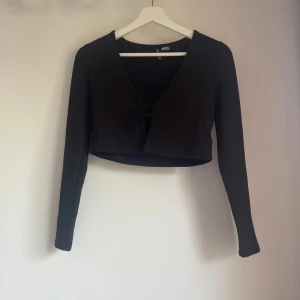 Svart croppad bolerotopp H&M - Svart ribbad bolerotopp från H&M Divided med lång ärm och croppad passform. Toppen har v-ringning och knyt framtill för en snygg detalj. Perfekt att slänga över ett linne eller klänning för en trendig layering-look.