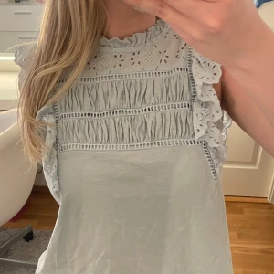 Ljusblå broderad blus med volang - Supersöt topp från Holly & Whyte. Broderade detaljer och volanger gör den perfekt till sommarens outfits, både till jeans och kjol. Lite trasig vid knappen bak (syns inte när man har den på) men går att sy eller byta. Använd 1 gång