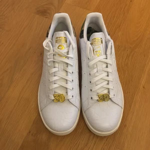 Adidas Stan Smith x BAPE sneakers - Unika oanvända Adidas Stan Smith x BAPE sneakers i vitt skinn med gulddetaljer och camo-mönstrad häl. Guldiga 30-detaljer på snörningen och extra camo-skosnören medföljer. Perfekta för dig som vill sticka ut med limited edition-sneakers.
