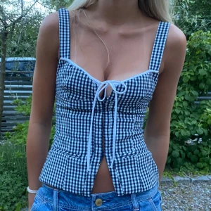 Rutig korsett-topp med knytning - Supersöt korsett-topp i svartvitt rutigt mönster med breda axelband och snygg knytning framtill. Figurnära passform och markerad midja, perfekt till jeans eller kjol. Toppens material känns lätt och somrigt.