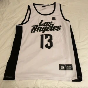 Vit och svart basketlinne FSBN - Snyggt basketlinne från FSBN i vitt med svarta detaljer och texten 'Los Angeles' samt siffran 13 på framsidan. Atletisk passform och tillverkat i polyester, perfekt för sport eller streetwear. Svarta paneler längs sidorna och broderad patch nertill.