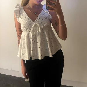 Vit blus med volang och knyt - Supersöt vit blus med broderade hål, volangdetaljer på axlarna och v-ringning. Toppen har knyt framtill och är ärmlös