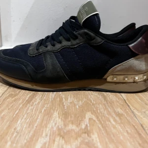 Valentino Garavani Rockrunner sneakers - Snygga Valentino Garavani Rockrunner sneakers i svart och mörkgrå mesh och mocka, med detaljer i vinrött och beige. Skorna har coola nitar på hälen och klassisk snörning. Perfekta för dig som vill ha en lyxig och unik sneaker med streetkänsla.