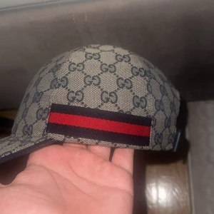 Gucci keps med GG-mönster och röd rand - Snygg keps från Gucci med klassiskt GG-monogram i grått och svart. Kepsen har en ikonisk röd och marinblå rand på sidan samt justerbart spänne i svart skinn baktill. Tillverkad i bomullsmix med exklusiv känsla och tydlig logga invändigt.