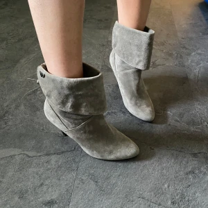 Grå mocka stövletter med rosett - Snygga grå stövletter i mocka med spetsig tå och hög klack. De har en bred, vikt skaftkant och en elegant metallrosett bak. Perfekta för dig som vill ha en stilren och trendig look.