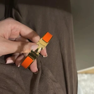 Guldarmband med orange detaljer - Snyggt armband i guldton med starkt orangea detaljer. Stelt och stilrent med klassisk H-formad låsning framtill. Perfekt accessoar för att ge din outfit en färgklick och exklusiv känsla.