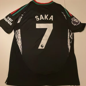 Arsenal Saka #7 bortatröja Adidas - Arsenal bortatröja replika med Saka #7, svart bas med röda och gröna detaljer på axlarna. Adidas-logga och Emirates Fly Better-tryck framtill, Premier League-märke på ärmen. Snygga vita mönster längs sidorna. Tillverkad i lätt funktionsmaterial. Endast handtvätt