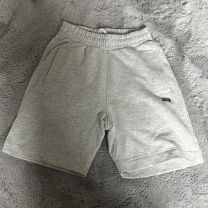 Grå sweatshorts från Yôke XS - Mysiga grå sweatshorts från Yôke med elastisk midja och snörning. Shortsen har två sidofickor och en bakficka, samt en liten svart logotyp på ena benet. Perfekta för chill dagar eller träning. Mjuk bomullskänsla och loose fit. Perfekt nu till sommaren! Hör av er vid eventuella funderingar och frågor! Nypris 550. 