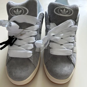 Grå adidas campus med mocka och vit sula - Snygga adidas campus i grå mocka med vita detaljer och klassiska tre ränder på sidan. Skorna har breda vita skosnören, gummisula i ljusbrun färg och adidas logga på plösen. Perfekta för dig som gillar stilrena och sportiga vibes.