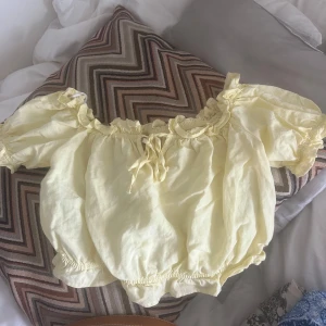 Gul offshoulder blus med volang - Supersöt ljusgul blus med offshoulder och puffiga korta ärmar. Toppen har volangdetaljer och knytning framtill, samt resår vid ärmslut och nederkant för en ballongig look. Perfekt för en somrig vibe!