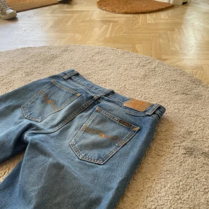 Blå jeans från Nudie Jeans - Säljer ett par klassiska blå jeans från Nudie Jeans med snygga kontrastsömmar och ikonisk brodering på bakfickorna. Jeansen har rak passform och normal midja, tillverkade i slitstarkt bomullstyg. Perfekta för en avslappnad och stilren look.
